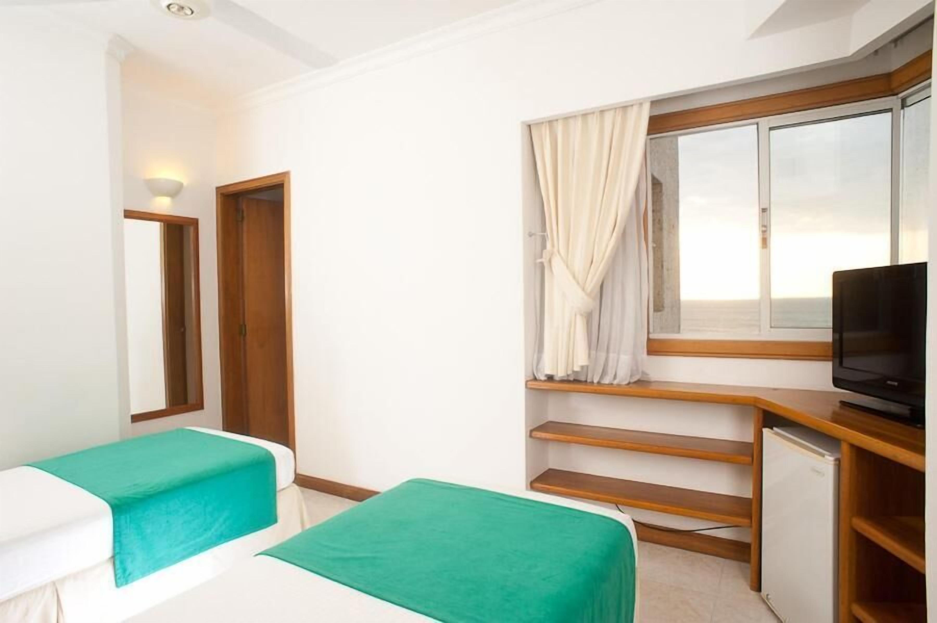 A hotel room with two beds, a television, a small refrigerator, and a window with curtains.
