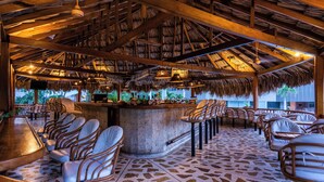 A bar area with a thatched roof, wooden beams, and stone tile flooring. There are bar stools and a bar counter with various items on it. - Hotel Irotama XXI (Santa Marta)