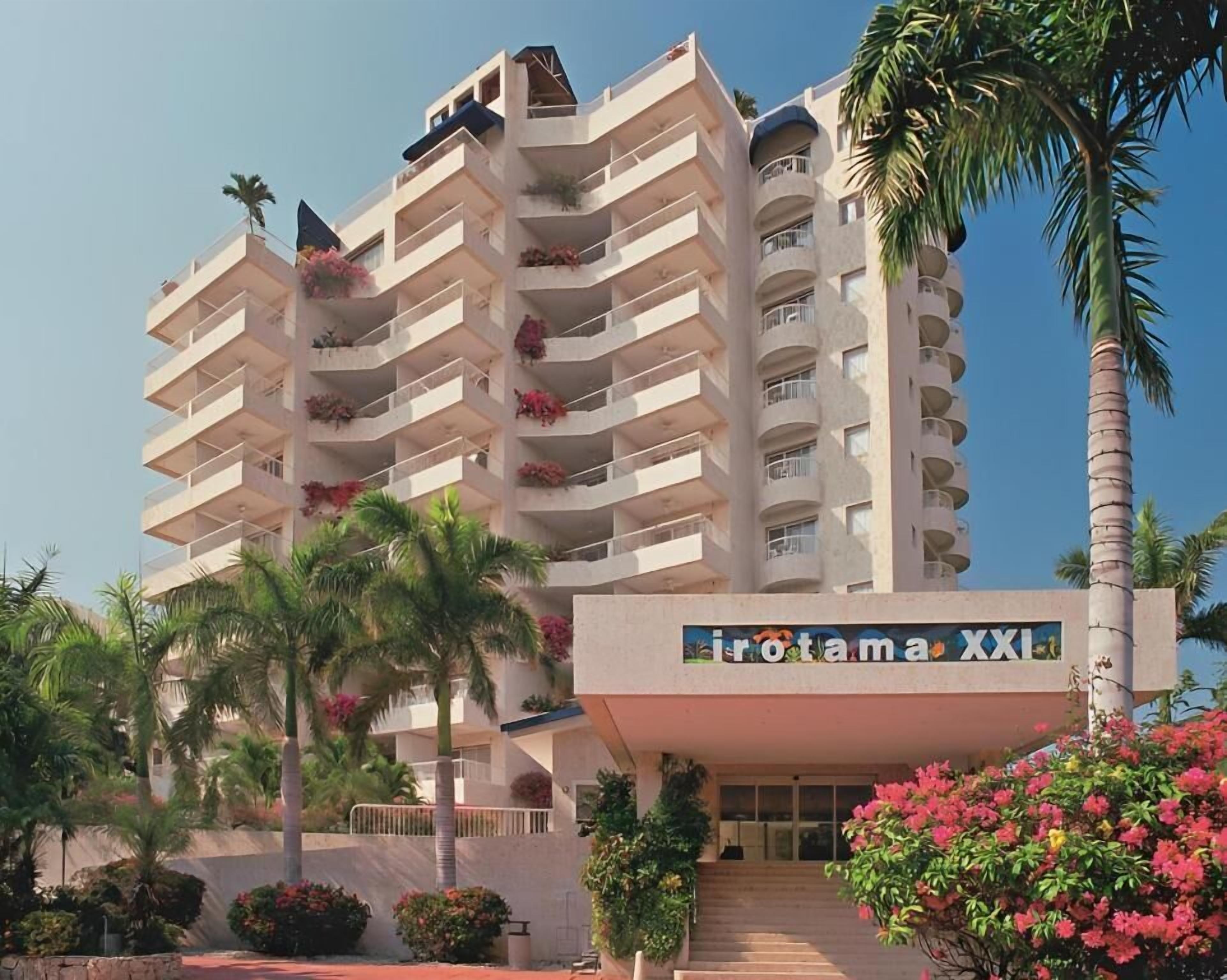 Un hôtel à plusieurs étages, doté de balcons ornés de fleurs, de palmiers et surplombant un ciel dégagé.