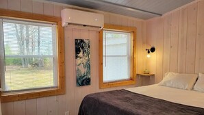Un dormitorio con cama, dos ventanas con persianas, aire acondicionado de pared, lámpara de noche y un cuadro en la pared.