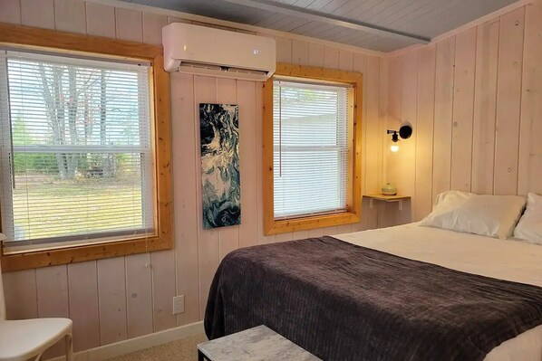 Un dormitorio con cama, dos ventanas con persianas, aire acondicionado de pared, lámpara de noche y un cuadro en la pared.