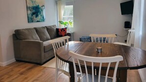 Una sala de estar con un sofá gris, una mesa de comedor de madera con sillas blancas, un cojín con forma de langosta roja y un cuadro en la pared.