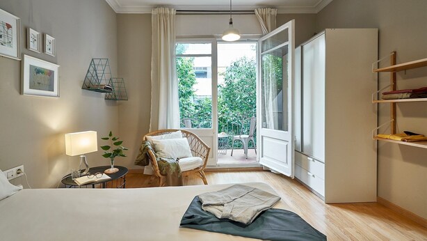 Ein Schlafzimmer mit Bett, Sessel, Tisch mit Lampe, Bücherregal und Blick ins Freie.