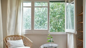Ein Zimmer mit einem Korbstuhl, einem kleinen runden Tisch mit einer Pflanze und einem Fenster mit Blick auf Grünflächen.