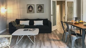 Ein Wohnzimmer mit einer schwarzen Couch, einem weißen Couchtisch und einem Essbereich mit Tisch und Stühlen. An der Wand hängen zwei gerahmte Bilder.
