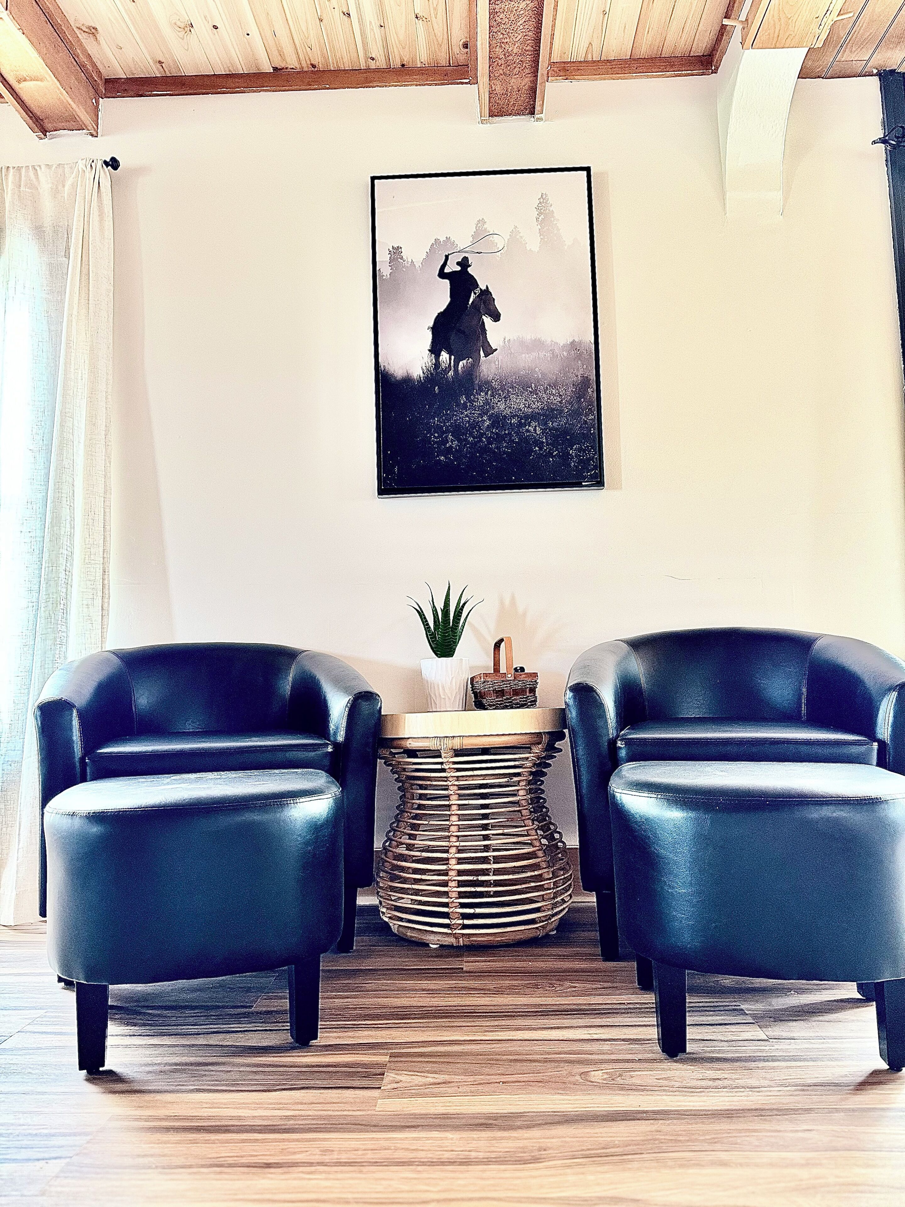 Ein Zimmer mit zwei schwarzen Ledersesseln, einem kleinen runden Tisch mit Flechtgeflecht und einem gerahmten Bild an der Wand.