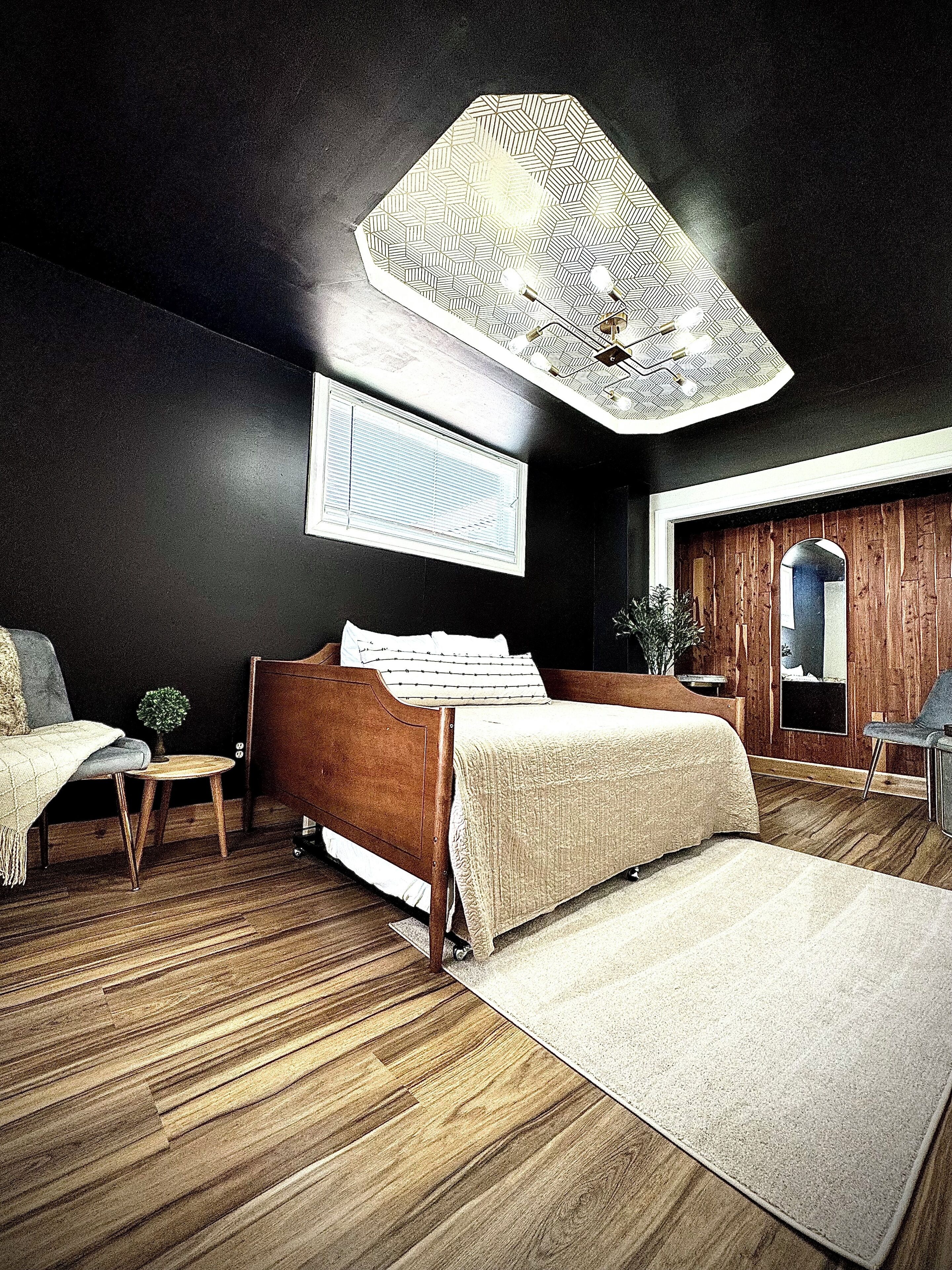 A bedroom with a wooden bed, a small bedside table, a chair, a large window, and a ceiling light fixture.