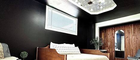 A bedroom with a wooden bed, a small bedside table, a chair, a large window, and a ceiling light fixture.