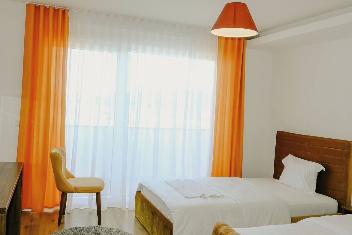 Une chambre d’hôtel avec deux lits, un bureau, une chaise et une grande fenêtre avec des rideaux légers.