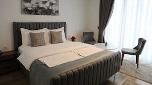 A hotel room with a large bed, two pillows, a bedside table with a lamp, a chair, and a painting on the wall. - Andoena Resort (Lipjan)