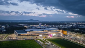 A modern building with a large glass facade, surrounded by parking areas and a landscaped green space. - Andoena Resort (Lipjan)
