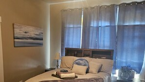 A bedroom with a bed, a nightstand, a lamp, a window with curtains, and a painting on the wall. - Central! Minutes to Magnolia, Baylor University, Cameron Park  Zoo and more.  (Waco)