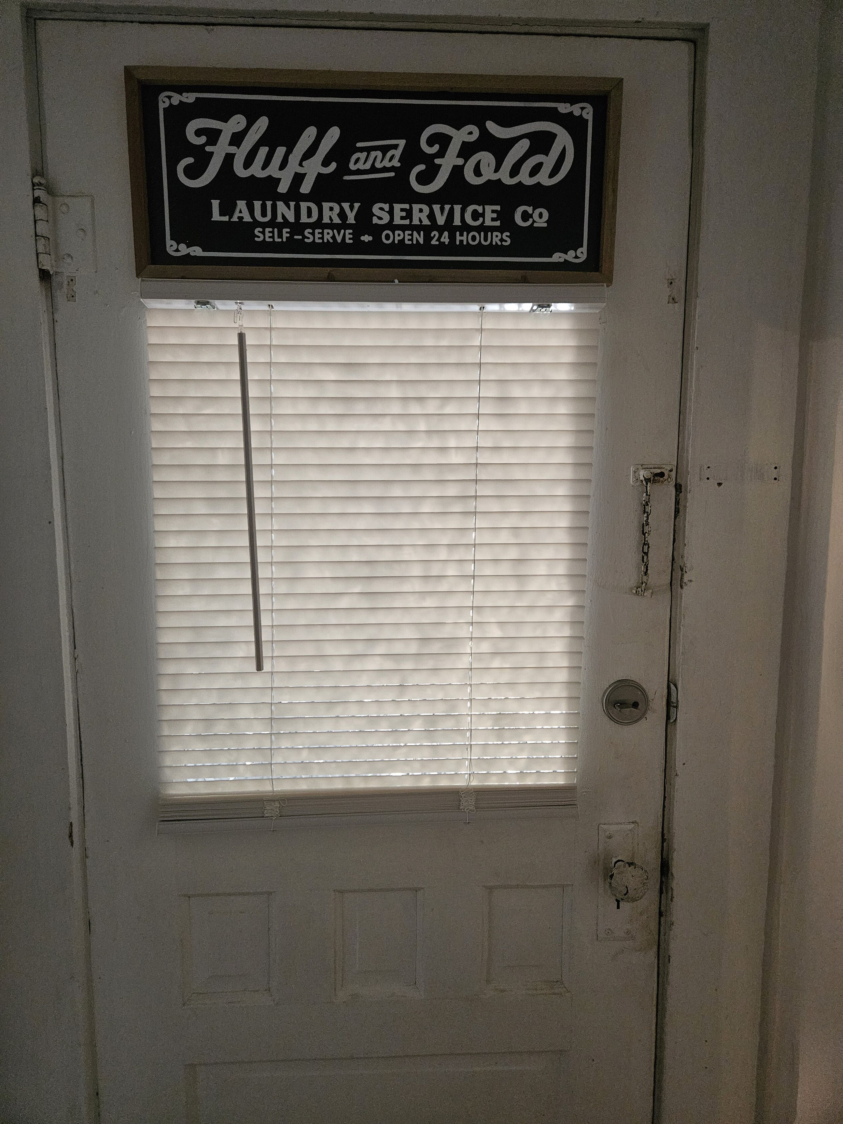 Una porta amb un rètol que diu 'Fluff and Fold Laundry Service Co. Autoservei Obert 24 Hores'.