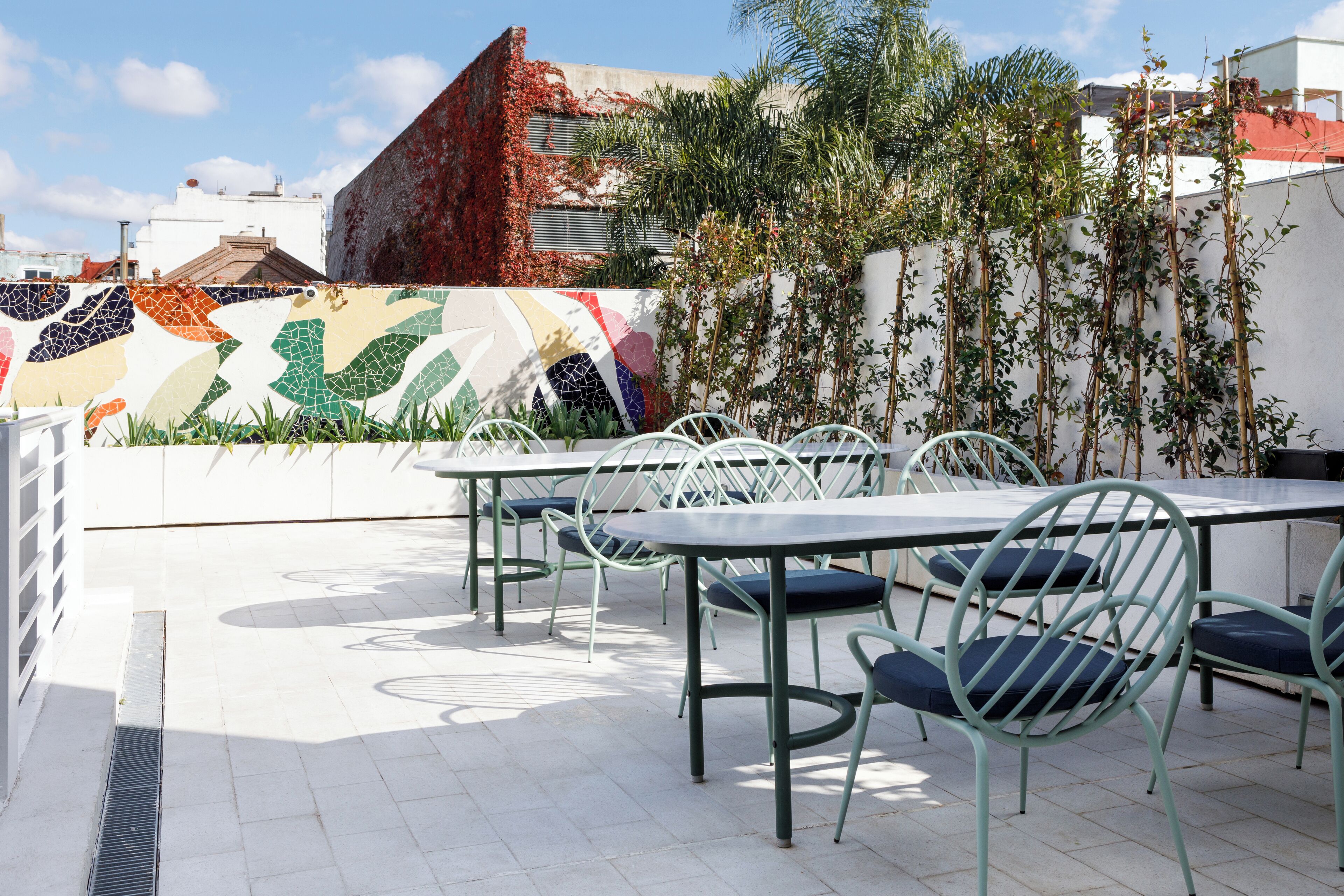 Un patio al aire libre con mesas y sillas de metal blanco, piso de baldosas y una pared decorativa con diseño de mosaico.