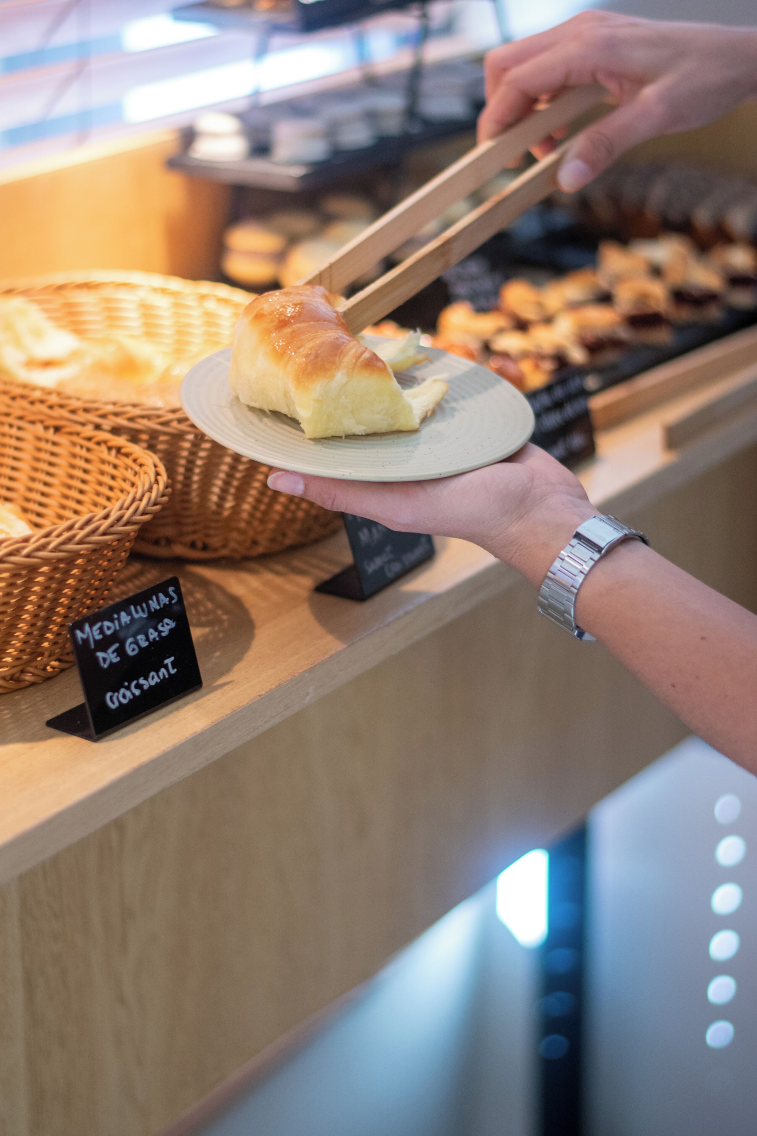 A hand holding a plate with a croissant, a basket of bread, and a sign indicating 'Medialunas de Graso Croissant'.