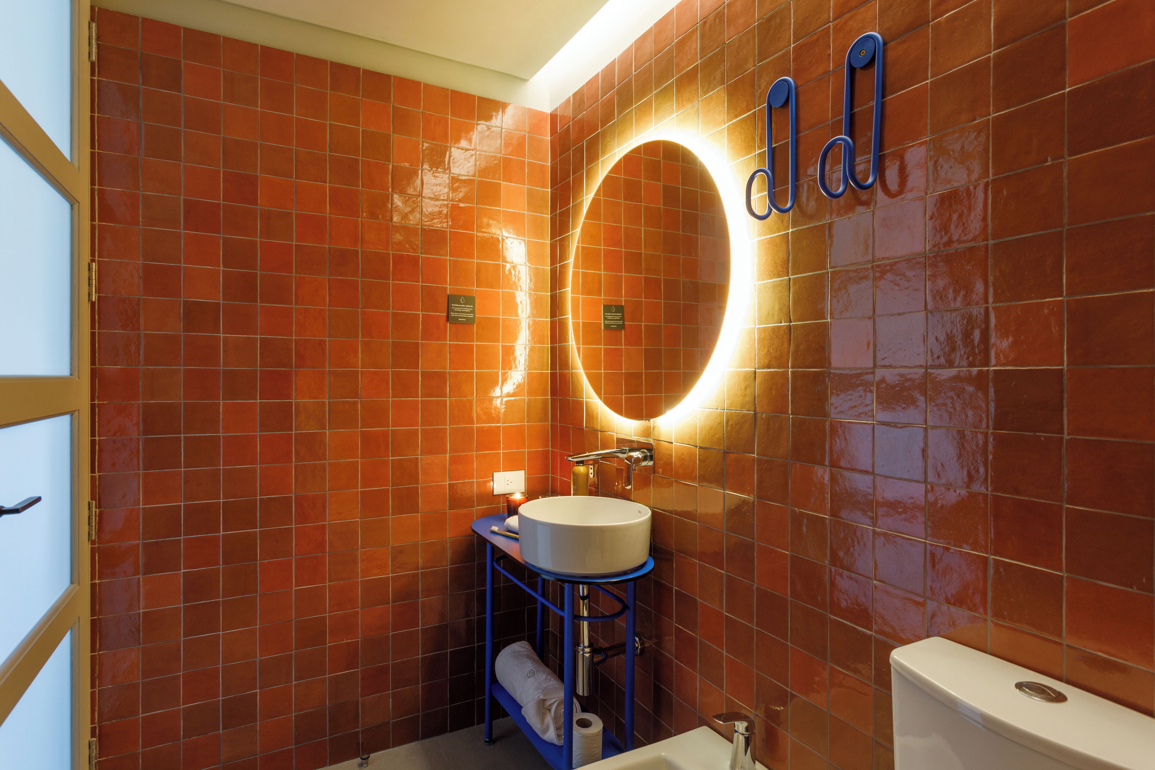 A bathroom with orange tiled walls, a round mirror, a white sink, and a blue vanity.