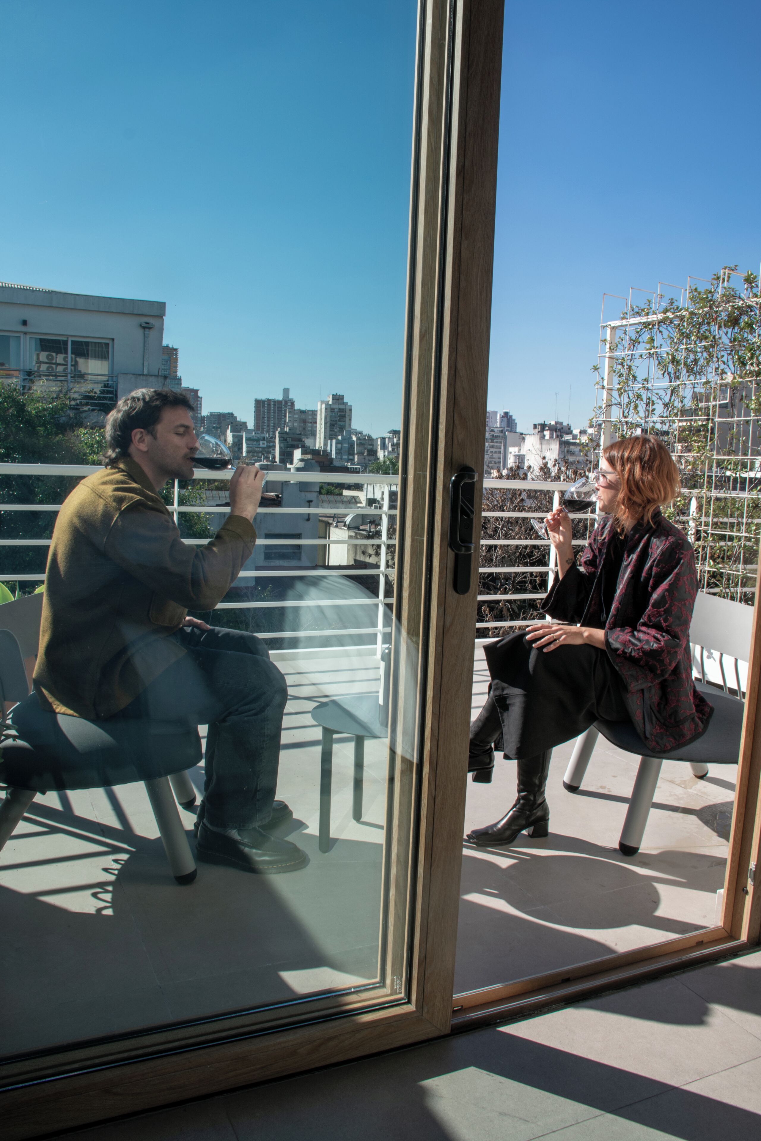Two people sitting on a balcony with a city view, a glass door, and a railing.