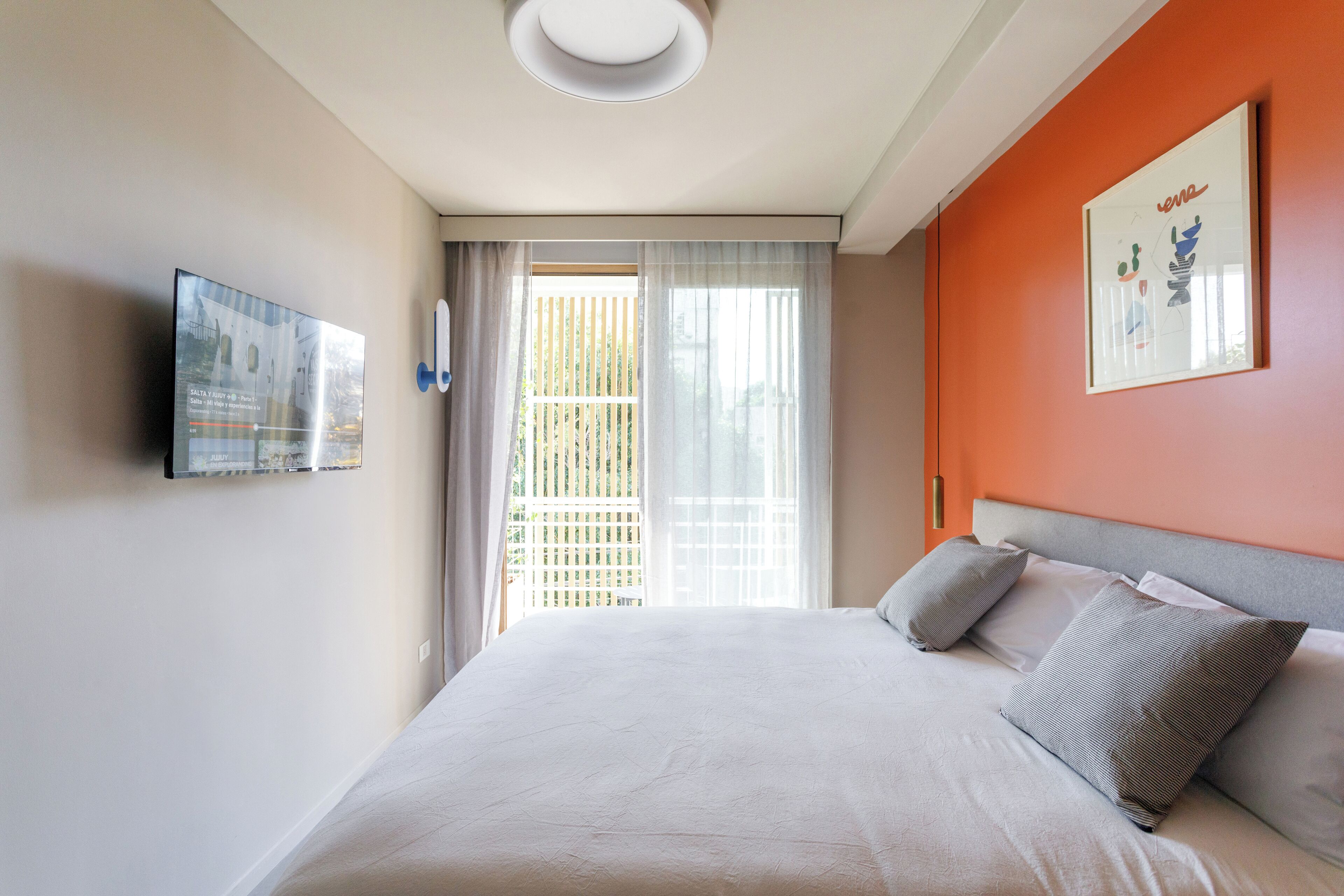 Une chambre à coucher avec un lit, un téléviseur fixé au mur et une fenêtre avec des rideaux.