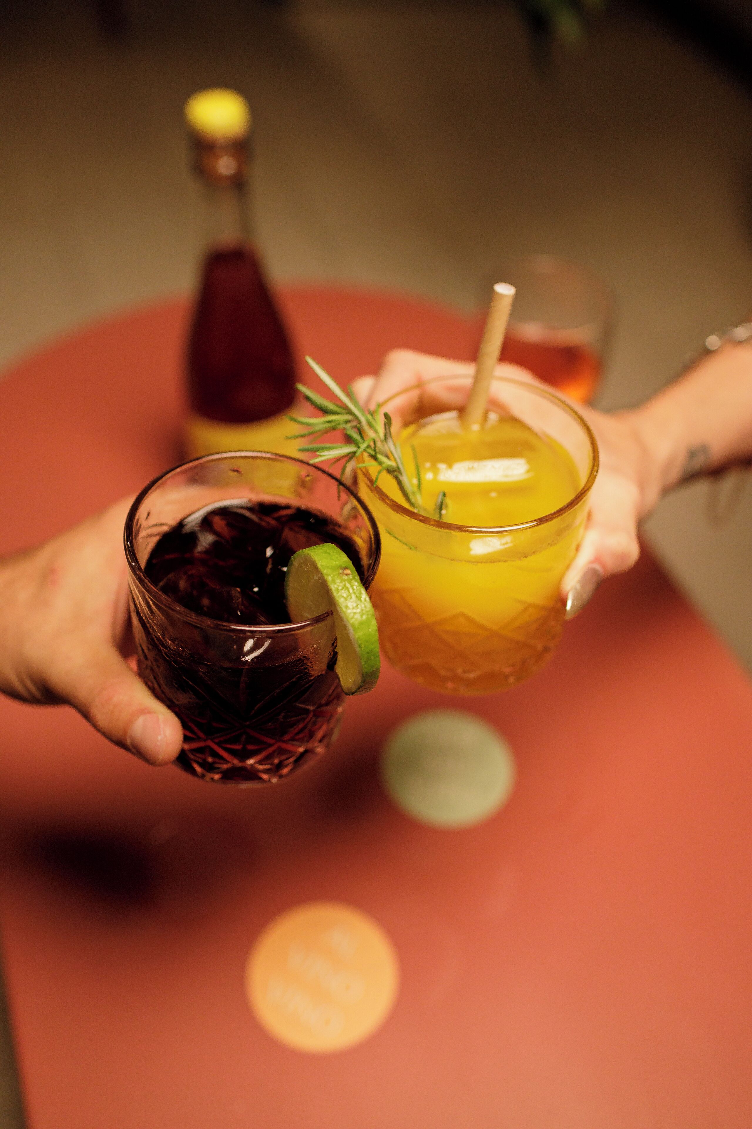 Two hands holding glasses of drinks with a bottle and a glass in the background.