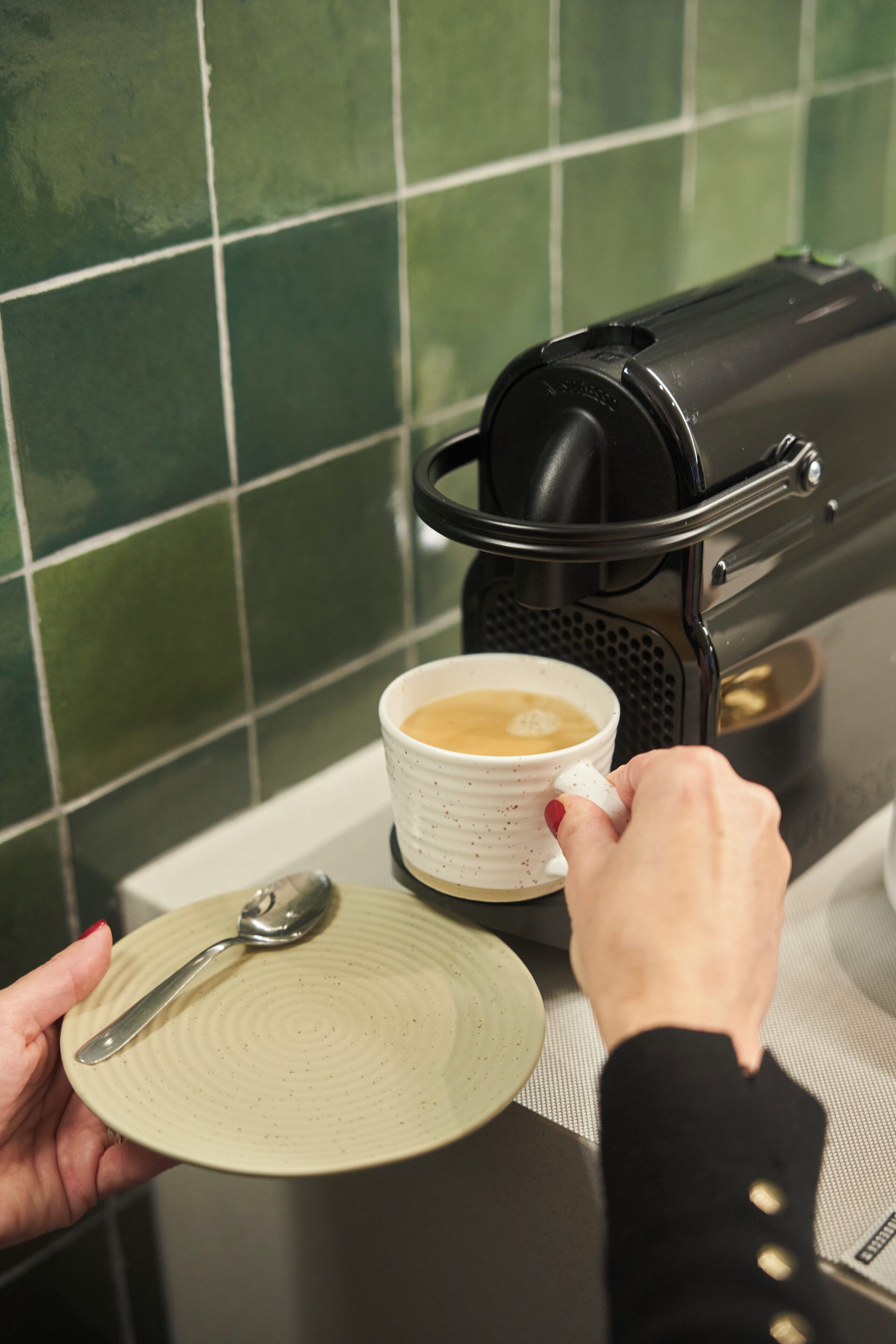 Una mano sosteniendo un plato con una cuchara, una taza de café y una cafetera.