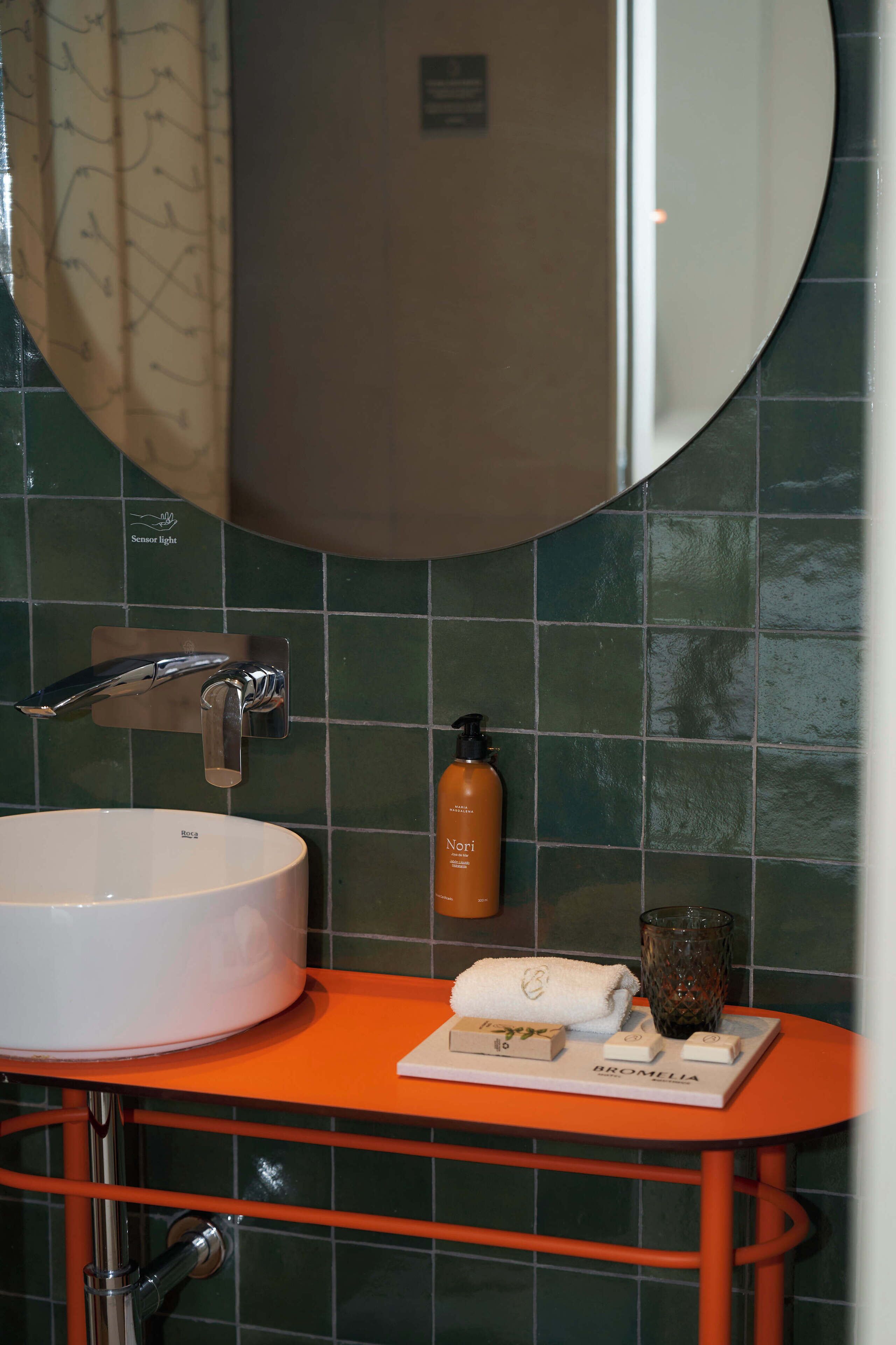 A bathroom with a white sink, chrome faucet, and a mirror above it. There is an orange countertop with toiletries and a glass.