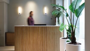 Een receptiebalie met verticale houten lamellen, twee wandlampen en een grote potplant aan de rechterkant.