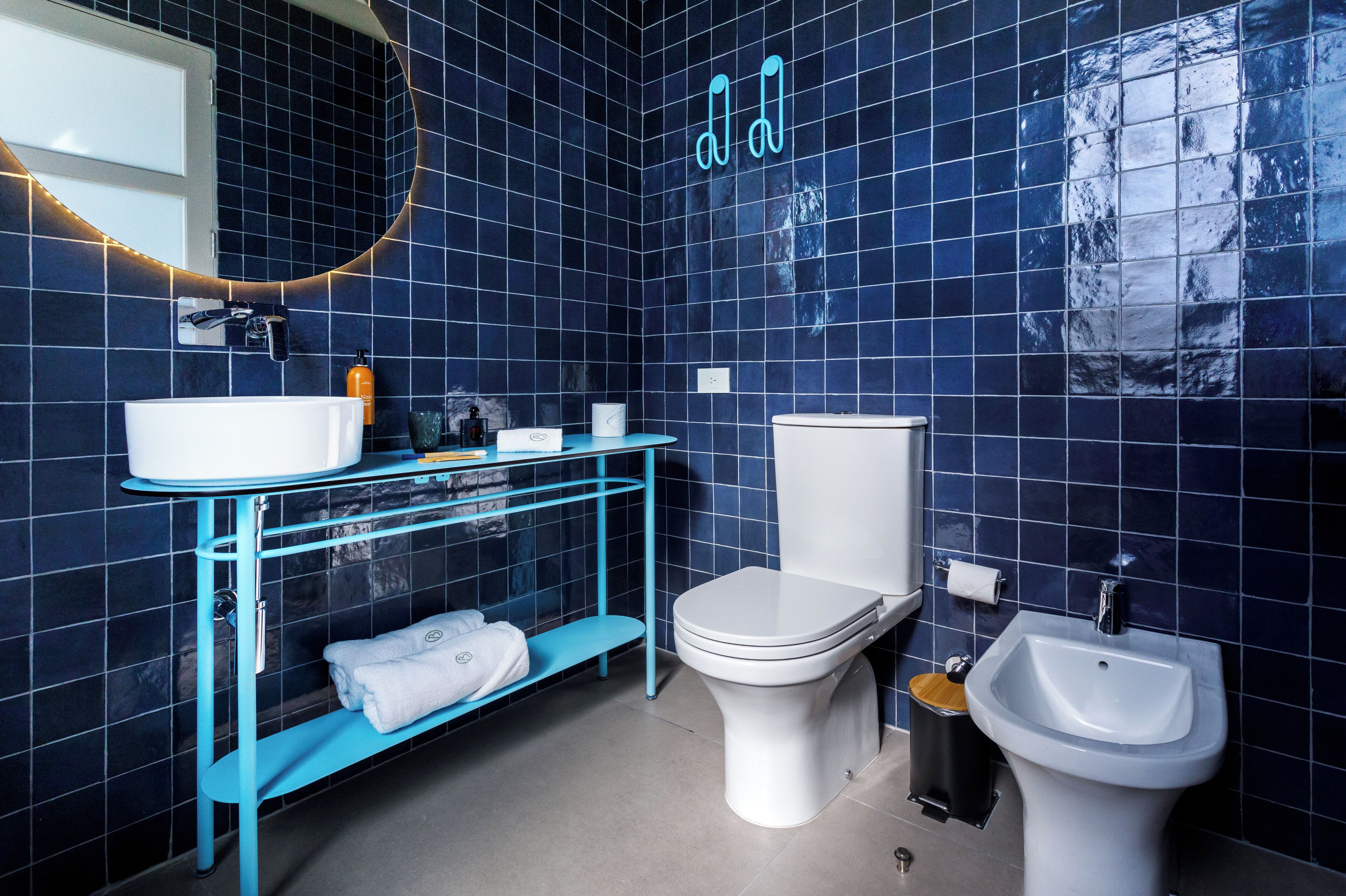 A modern bathroom with a white toilet, a white sink, and a bidet. There is a blue shelf with towels and toiletries.