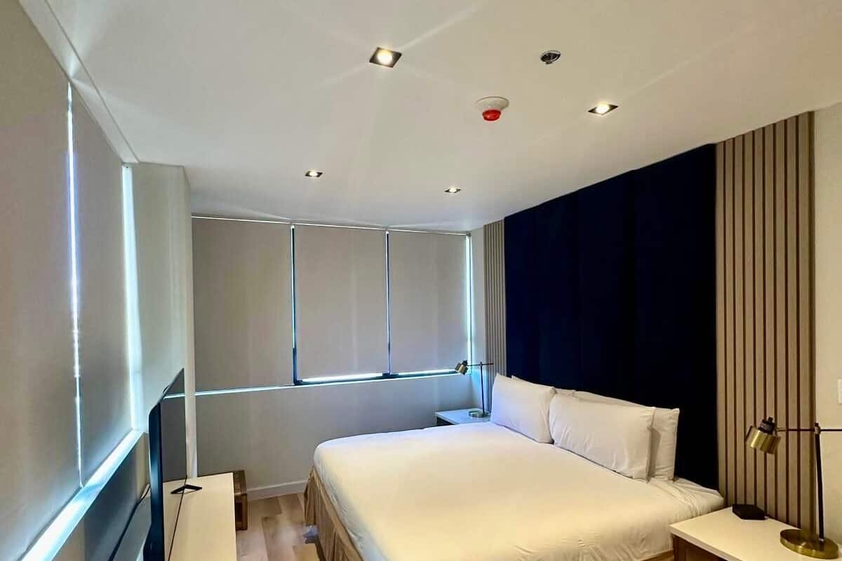 Een moderne slaapkamer met een groot bed, nachtkastjes, een televisie en een raam met jaloezieën.