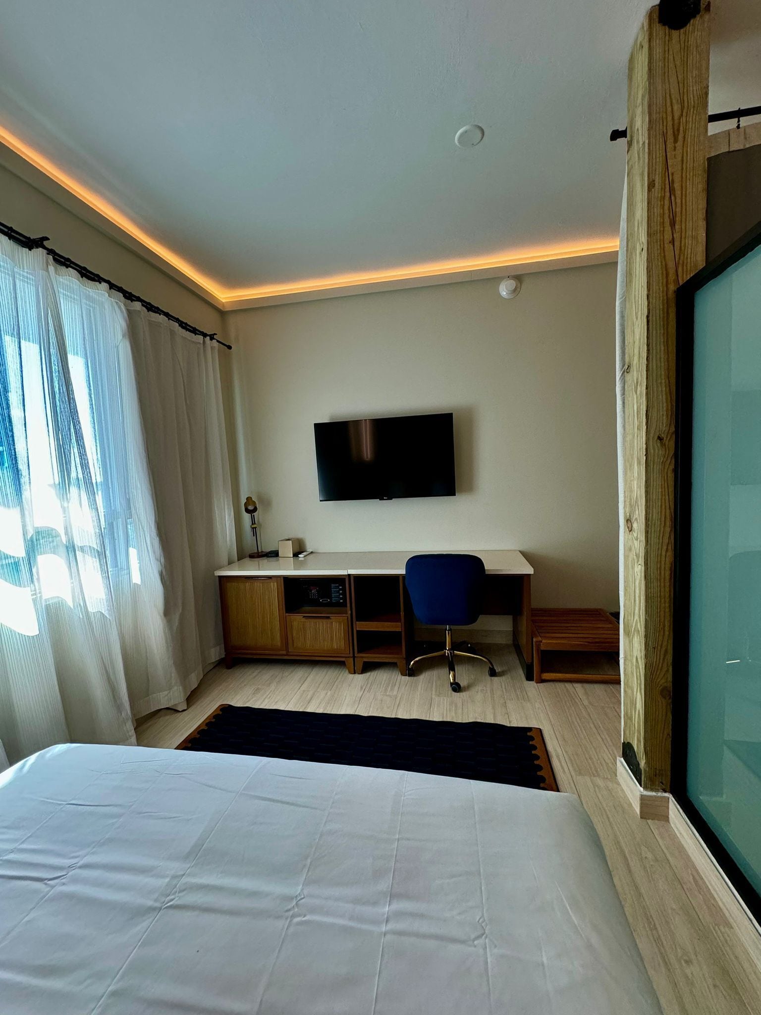 Ein modernes Hotelzimmer mit einem großen Bett, einem Schreibtisch mit Stuhl, einem Fernseher und einem großen Fenster mit Vorhängen.