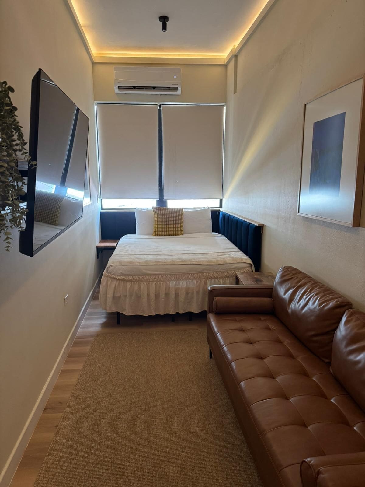 A narrow hotel room with a bed, a brown leather sofa, a large mirror, and an air conditioning unit.