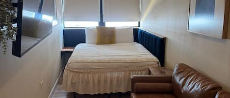A narrow hotel room with a bed, a brown leather sofa, a large mirror, and an air conditioning unit.