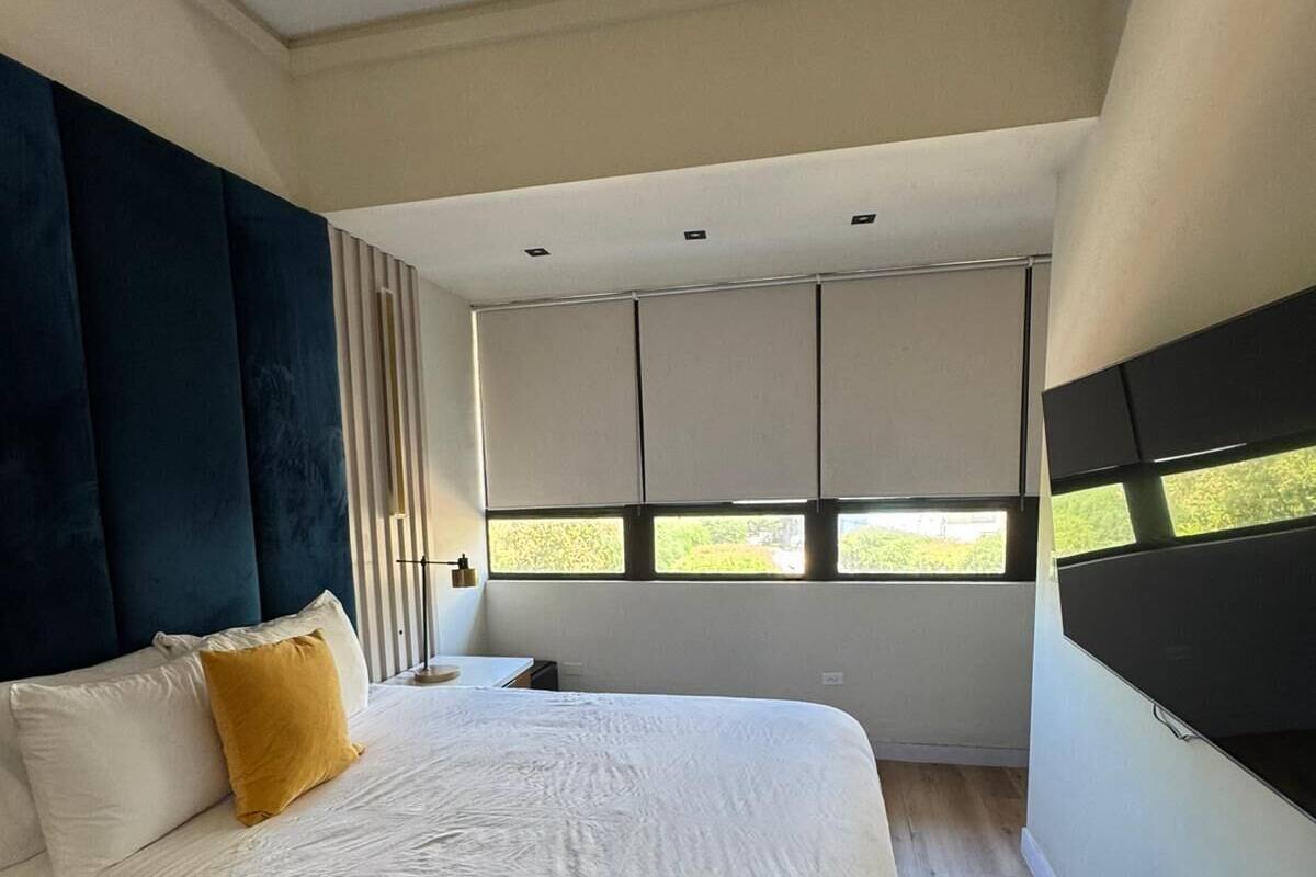 Een slaapkamer met een bed, een nachtkastje, een raam met zonwering en een aan de muur bevestigde televisie.