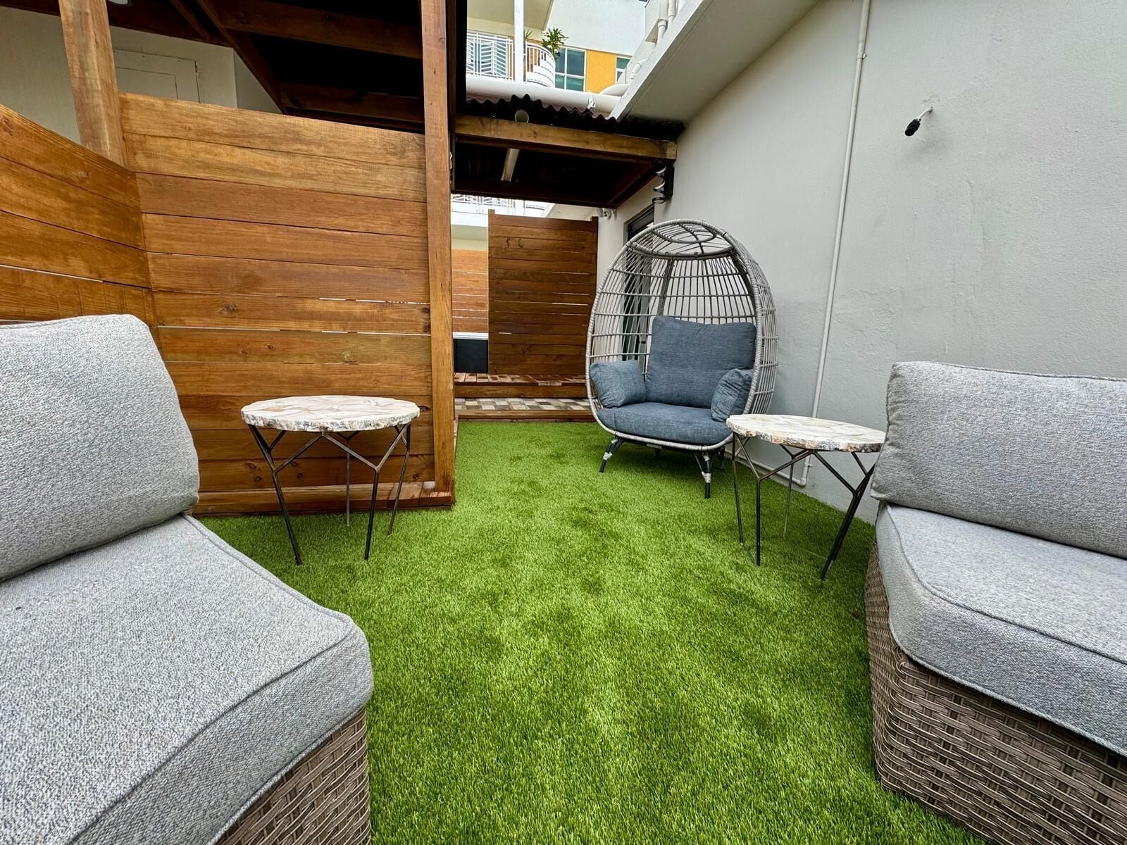 Un pequeño espacio al aire libre con dos sofás grises, un sillón de mimbre y una pequeña mesa redonda, todo sobre césped artificial.