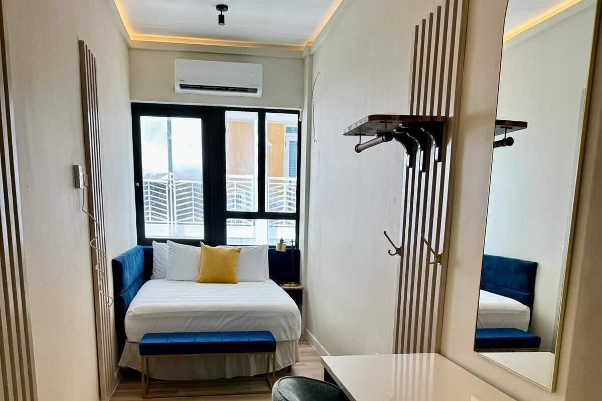 Een moderne hotelkamer met een bed, een bankje, een kaptafel met spiegel en uitzicht op gebouwen door het raam.