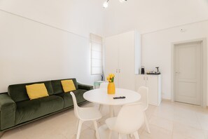 Una sala de estar moderna con un sofá verde, sillas blancas, una mesita redonda y un jarrón con flores.