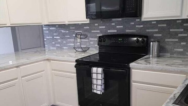 A kitchen with white cabinets, a black stove, a microwave, and a tiled backsplash. - Cute And Cozy Hostel (Houston)