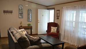 Una sala de estar con sofá, sillón y mesa de centro. Se ve una puerta, una ventana con cortinas y adornos en la pared.