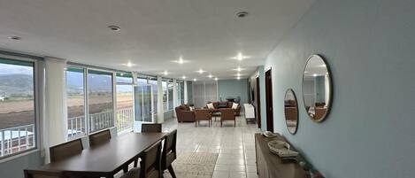 A spacious dining area with a long table and chairs, a sofa set, and a large mirror on the wall.