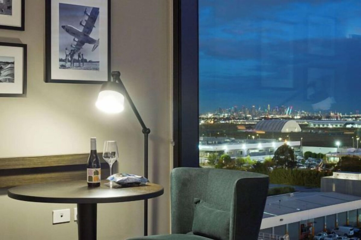 A room with a view of a cityscape at night, a table with a bottle and glasses, and a chair.