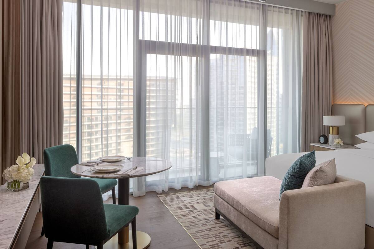 Een hotelkamer met een bed, een bureau, een stoel en uitzicht op de stad door grote ramen.