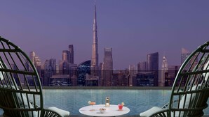 Una piscina en la azotea con dos sillas de mimbre y una mesa, con vistas a un paisaje urbano con rascacielos modernos, incluyendo una torre destacada.