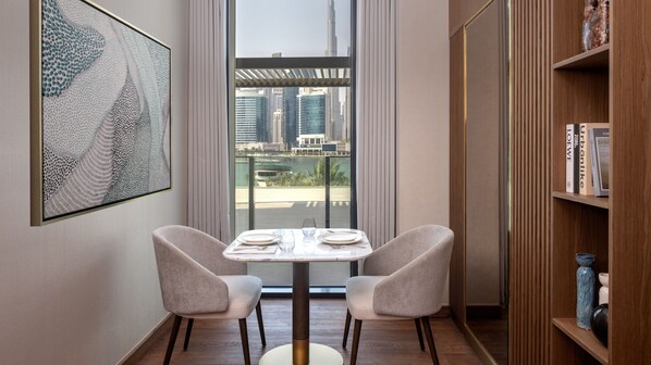 A modern room with a dining table set for two, a bookshelf, and a large window with a city view.