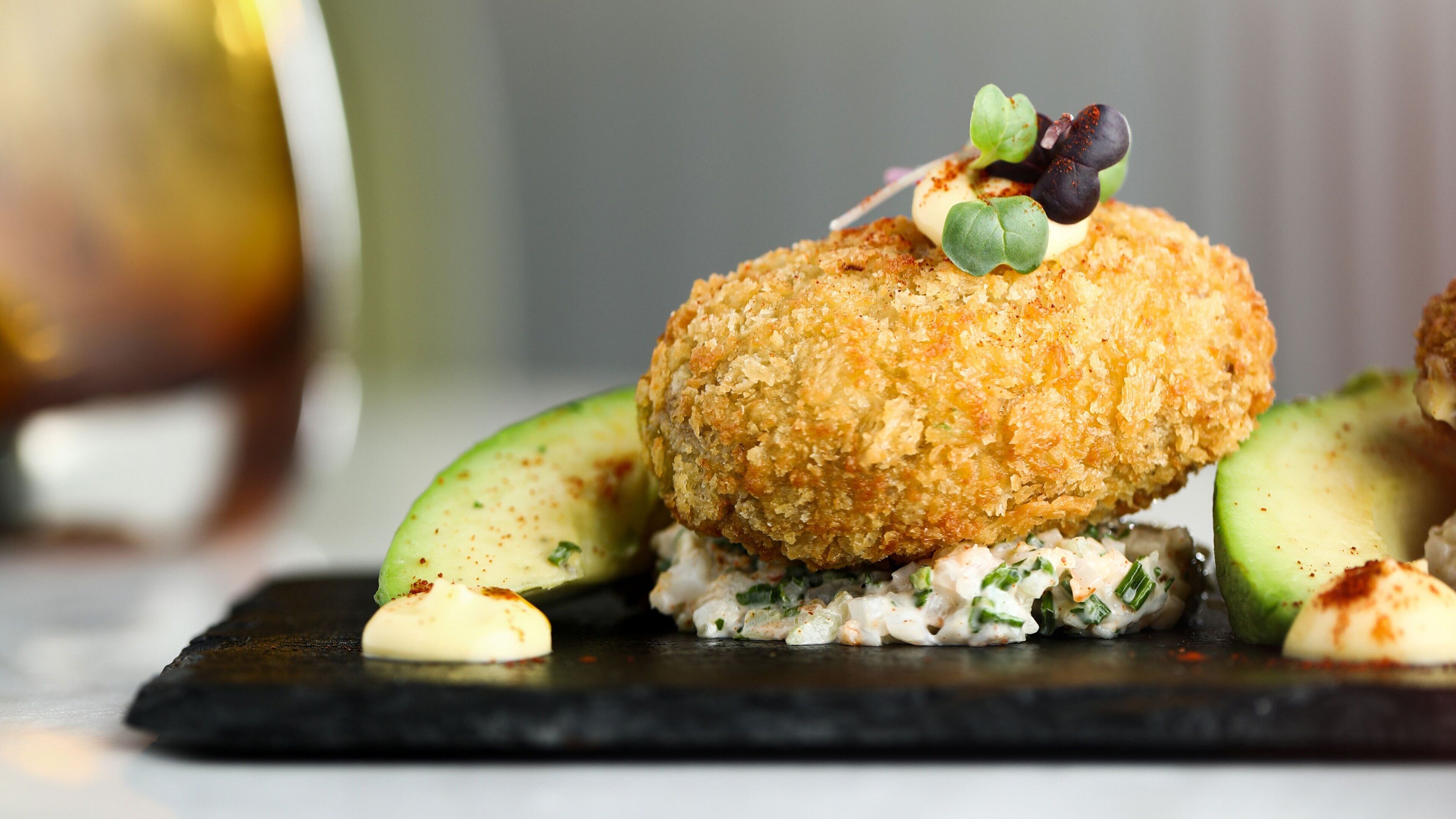 Un plat composé d'un aliment pané, de tranches d'avocat et d'une sauce crémeuse, le tout présenté sur une assiette foncée.