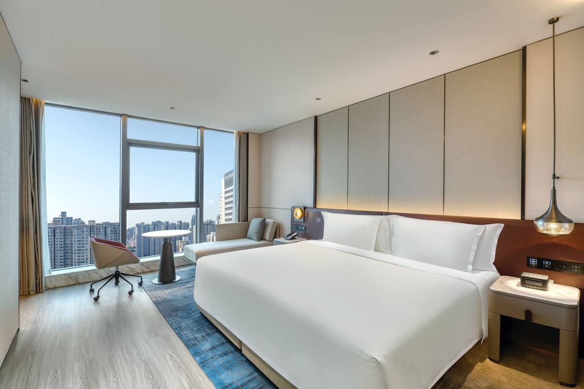 A modern hotel room with a large bed, a desk, and a chair. There is a large window offering a city view.