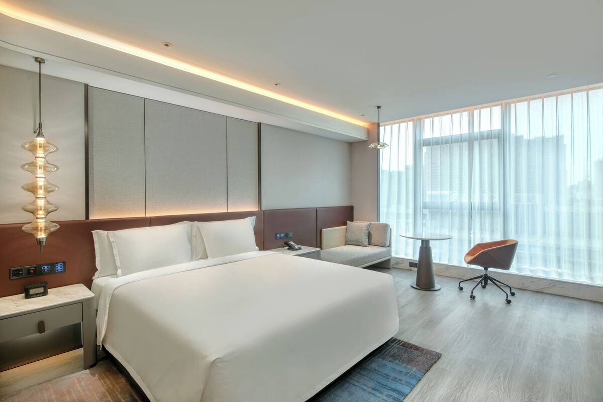 A modern hotel room with a large bed, a desk, a chair, and a large window with curtains.