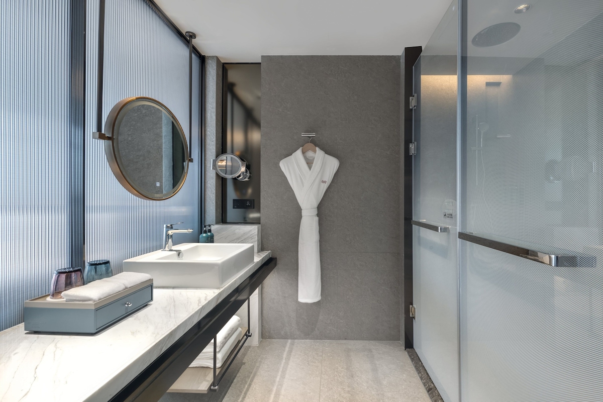 A modern bathroom with a dual sink vanity, a round mirror, a robe hanging on the wall, and a shower enclosure.