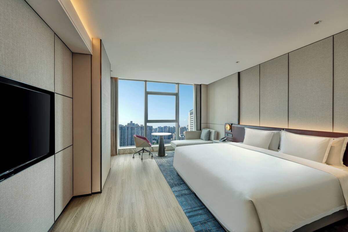 A modern hotel room with a large bed, a flat-screen TV, a small table, and a chair.