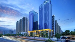Um hotel moderno com fachada de vidro marcante, vários edifícios altos ao fundo e uma rua urbana bem cuidada em primeiro plano.
