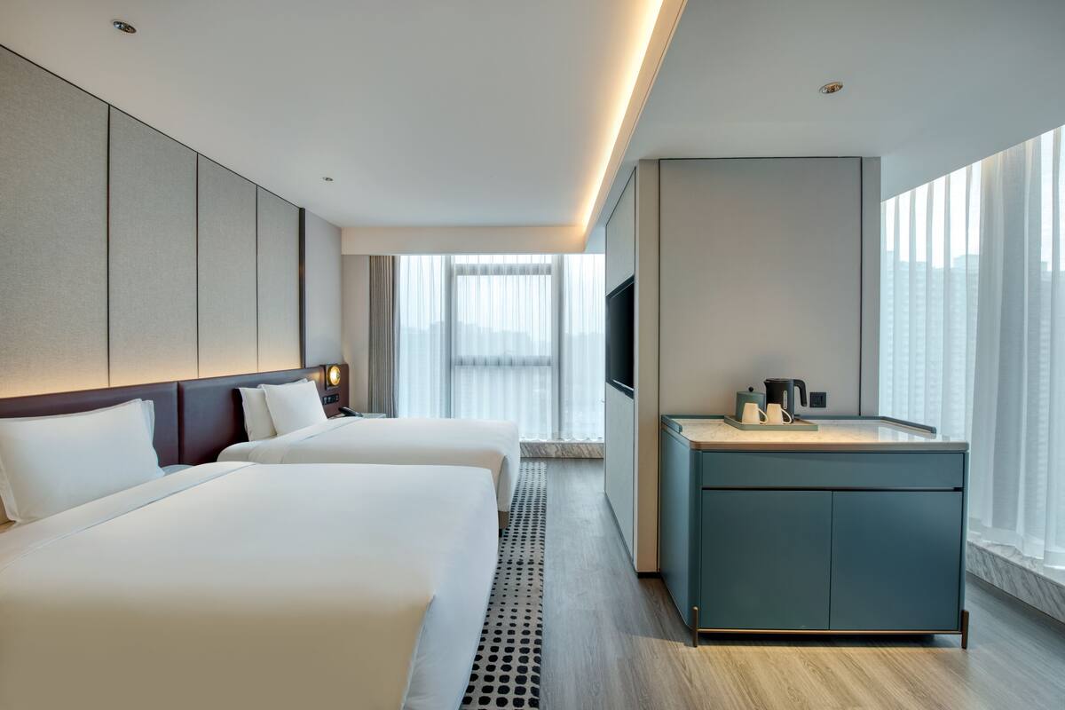A hotel room with two beds, a minibar, a TV, and a large window with a city view.
