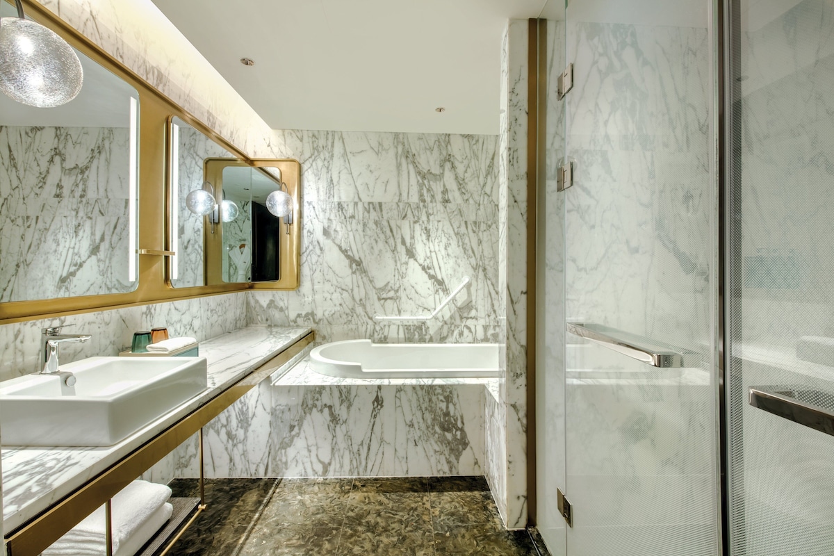 A modern bathroom with marble walls, a large mirror, two sinks, a bathtub, and a glass shower enclosure.